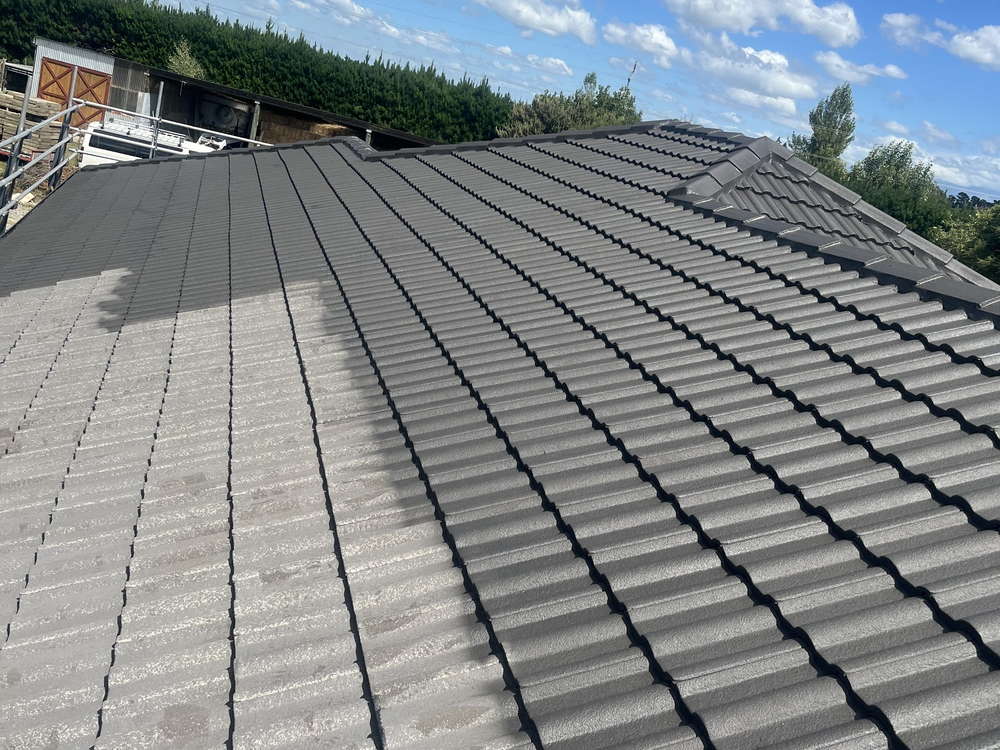 Concrete tile roof in Darfield half way through painting: bare grey tiles on the left, new charcoal coating on the right