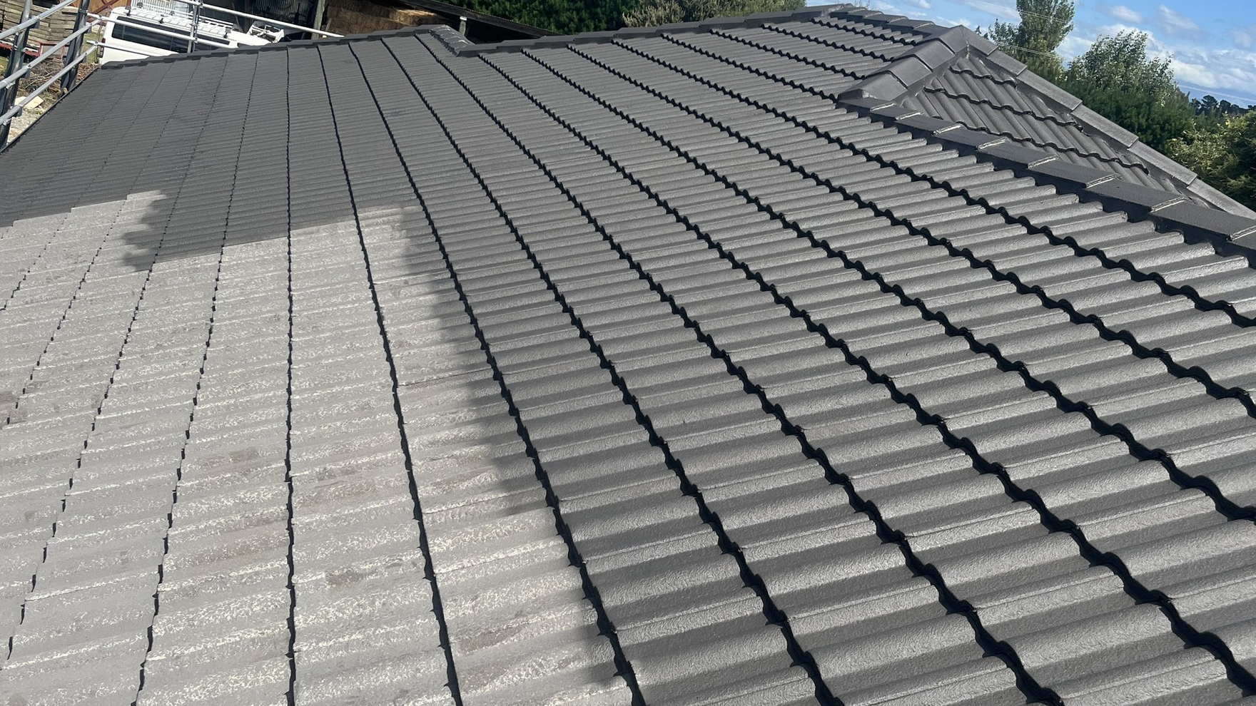 Concrete tile roof in Darfield part way through being painted, the left side still bare grey and the right side coated in charcoal